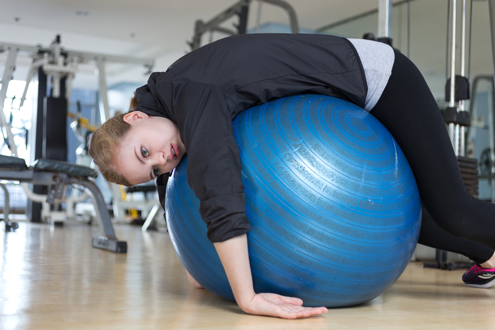 Young woman flopped over a blue yoga ball in a gym exhausted.